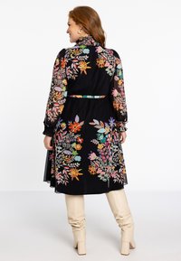 Schwarz mit floralen Stickereien versehenes Kleid mit langen, durchsichtigen Ärmeln, tailliertem Bund und einem rüschenden Kragen. Kombiniert mit hellbeigen kniehohen Stiefeln.
