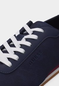 Gros plan sur une sneaker bleu marine avec des lacets blancs, une texture en suède et un logo "GUESS" embossé sur le côté.