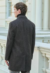 Mörkgrå ullkappa med texturerad yta, enkelknäppt design, slagkrage och cuffs med tre knappar, sedd bakifrån.