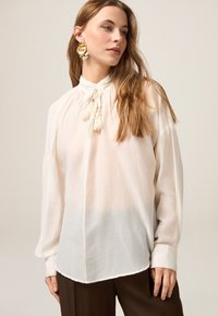 Blusa blanca, ligera, de manga larga con cuello alto, que presenta un cierre con lazo y detalles plisados. Textura suave, ajuste relajado.
