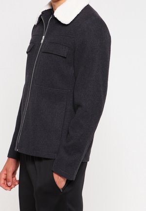 Homme portant une veste zippée gris foncé avec un col en polaire blanche et des poches poitrine, debout avec une main dans la poche du pantalon.