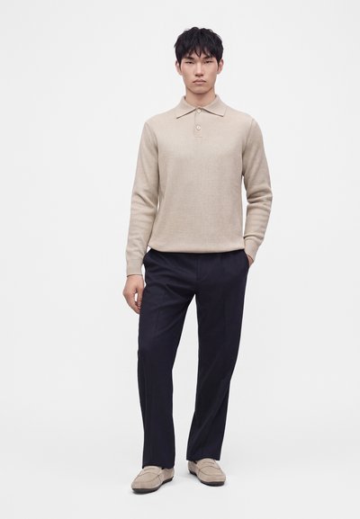 Homme debout portant un polo beige à manches longues, un pantalon bleu marine foncé et des mocassins beiges sur un fond clair uni.