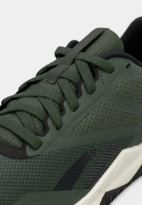 Scarpa da atletica verde con tomaia sintetica testurizzata, sezioni in rete, lacci piatti e accenti neri. La suola ha un design angolare con base bianca.