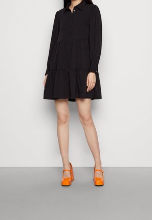 Kobieta ubrana w czarną, zapinaną na guziki sukienkę z długim rękawem, w jaskrawo pomarańczowe buty Mary Jane na grubym obcasie, stojąca na jednolitym tle.