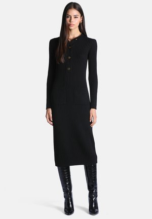 Femme aux longs cheveux foncés porte une robe noire ajustée côtelée avec des boutons dorés et des bottes en cuir verni noir montantes jusqu'aux genoux.