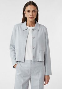 Lichtgrijze cropped jack met een kraag, knopen aan de voorkant en twee zijzakken; gedragen over een wit top met bijpassende broek.