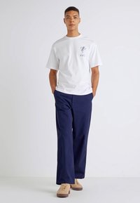 White cotton t-shirt with short sleeves and small graphic print, paired with dark navy wide-leg pants, and light tan lace-up sneakers.
