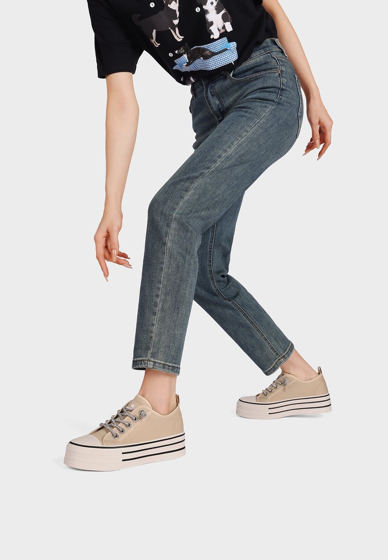 Persona con jeans azules y zapatillas plataforma beige dando un paso hacia adelante sobre un fondo blanco, con una camiseta gráfica negra parcialmente visible.