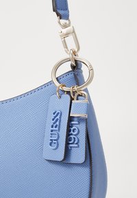 Bolsa sintética azul com superfície texturizada, acabamentos em prata e duas etiquetas gravadas com "GUESS" e "1981" presas a um anel.