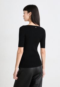 Črn rebrast srajc s kratkimi rokavi, oprijet kroj, ovalen izrez in majhen srebrn detajl na hrbtni strani. Nosijo ga z črnimi usnjenimi hlačami.