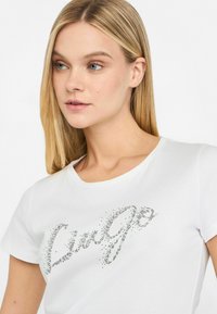 Hvid bomulds T-shirt med et funklende sølv "Live Go" grafik lavet af små rhinsten; korte ærmer, rund hals design.