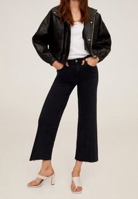 Schwarze Lederjacke mit kurzem Schnitt, weißes T-Shirt, hoch taillierte schwarze Jeans mit ausgestellter Silhouette und weiße Sandalen mit hohem Absatz.