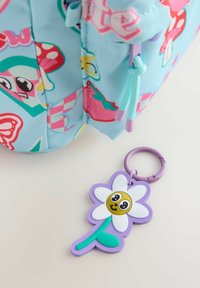 Detail eines mehrfarbigen Rucksacks mit himmelblauem Stoff, der lustige Muster aufweist. Ein lila, blumenförmiger Schlüsselanhänger mit einem lächelnden Gesicht hängt darunter.