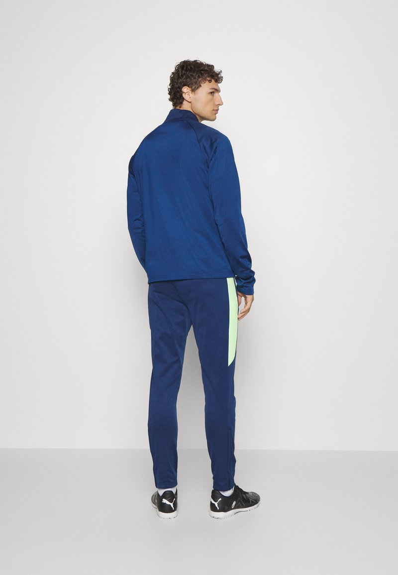 Navy sportjack met opstaande kraag en bijpassende broek met een lichtgroene zijstreep. Gedragen met zwarte sportschoenen.