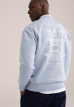 Uomo di profilo che indossa una felpa azzurra con uno schizzo bianco di un edificio e il testo "Downtown Social Club Bedford Avenue New York"