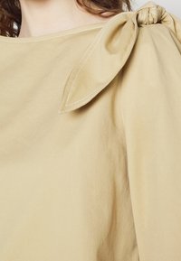 Haut en tissu beige avec un col bateau présentant un grand nœud cousu sur l'épaule, porté par une personne aux cheveux bruns.