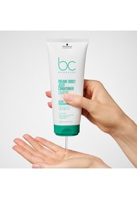 Tube blanc avec un capuchon bleu sarcelle distribuant un après-shampoing gelée claire dans une main. Le texte en bleu sarcelle et noir indique le nom du produit et son usage.
