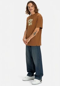 Hombre con cabello hasta los hombros y bigote, vestido con camiseta gráfica marrón, vaqueros azules holgados, zapatillas blancas, y mostrando un tatuaje de tiburón en el antebrazo.