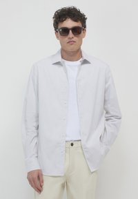 Camicia bianca a righe con colletto a punte, indossata aperta su una maglietta bianca, abbinata a pantaloni beige e occhiali da sole scuri.