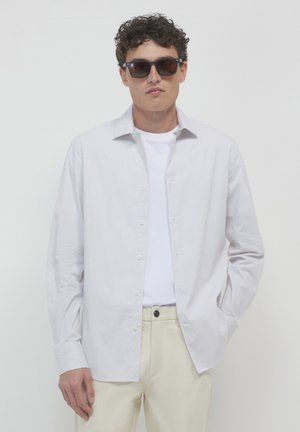 Chemise à rayures blanches avec un col évasé, portée ouverte sur un t-shirt blanc, assortie à un pantalon beige et des lunettes de soleil sombres.