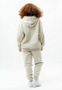 Personne aux cheveux bouclés de dos, portant un sweat à capuche beige, un pantalon de jogging assorti et des baskets blanches, debout sur un fond blanc.