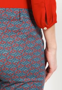 Modrá látka s červeným a růžovým vířícím vzorem, s postranní kapsou a knoflíkovým detailem na rukávu červeného topu.