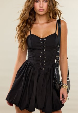 Femme aux longs cheveux ondulés portant une robe corset noire à lacets et tenant un sac en cuir noir avec des bracelets dorés au poignet.