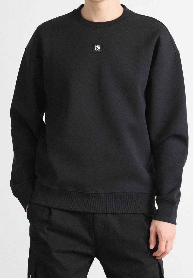 Personne portant un sweat-shirt noir uni avec un petit logo carré inscrit "HU GO" sur la poitrine, associé à un pantalon noir.