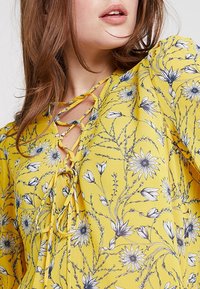 Blouse fleurie jaune avec un imprimé de fleurs bleues, un décolleté à lacets et un tissu doux et léger. Design élégant et coupe ample.