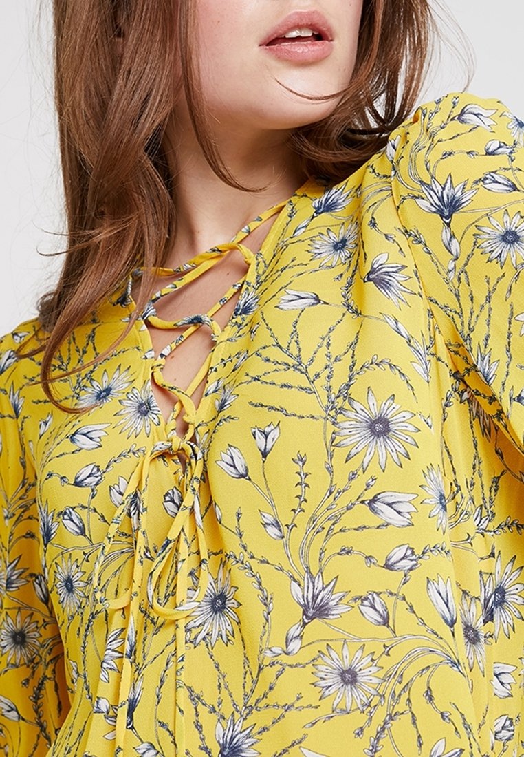 Blouse fleurie jaune avec un imprimé de fleurs bleues, un décolleté à lacets et un tissu doux et léger. Design élégant et coupe ample.