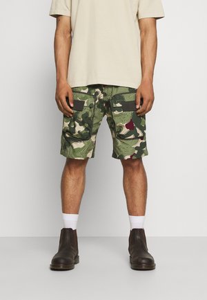 Homme portant un short cargo à motifs verts, un t-shirt beige, des chaussettes blanches et des bottes de cheville marron foncé, se tenant devant un fond uni.
