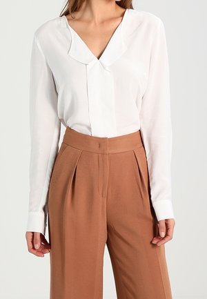 Femme porte un chemisier blanc à manches longues avec un col unique et un pantalon marron taille haute à jambes larges sur un fond uni.