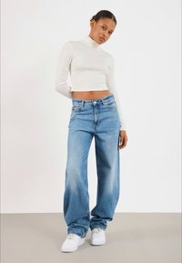 Woman wearing white long-sleeve cropped turtleneck, loose light blue jeans, and white sneakers, standing against plain light background.