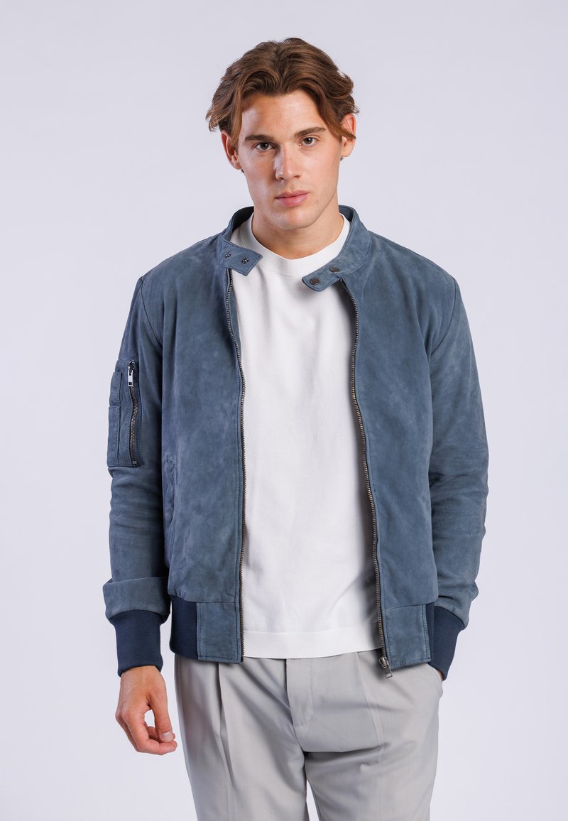Chaqueta bomber de ante azul con cremallera frontal, puños acanalados y un bolsillo con cremallera en la manga. Llevada sobre una camiseta blanca de cuello redondo y pantalones claros.