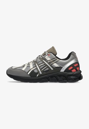 Zapatilla deportiva gris con parte superior de malla, acentos plateados reflectantes, suela negra y tela texturizada. Presenta un sistema de cordones flexible y un cuello acolchado.