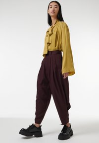 Blusa mostaza con escote volante, combinada con pantalones burdeos de talle alto. Zapatillas negras con suela gruesa completan el atuendo.