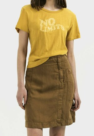 Personne portant un t-shirt jaune moutarde avec le texte "NO LIMITS" et une jupe marron jusqu'aux genoux avec de grandes poches.
