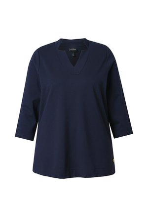 Tmavě modrý top s výstřihem do V a tříčtvrtečními rukávy, vyrobený z hladké látky; má malý emblém na spodní straně.