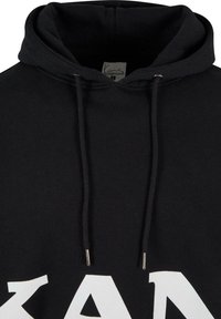 Hoodie noir avec cordons de serrage et grandes lettres blanches audacieuses "KAN" imprimées sur le devant, avec une étiquette de cou visible à l'intérieur de la capuche.
