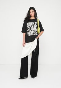 Μαύρο oversized t-shirt με γραφικό "MAKE SOME NOISE", λευκή λεπτομέρεια draped, μαύρο παντελόνι φαρδιάς γραμμής και πράσινη τσάντα χιαστί.