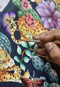 Bunt gefärbtes Leder mit detaillierten Blumen- und Tiermustern. Eine Hand mit einem Pinsel fügt dem Kunstwerk auf einem dunklen Hintergrund feine Akzente hinzu.