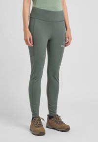 Groene, rekbare legging met een hoge tailleband en zijzak; gecombineerd met bruine wandelschoenen met veters en gestructureerde zolen.