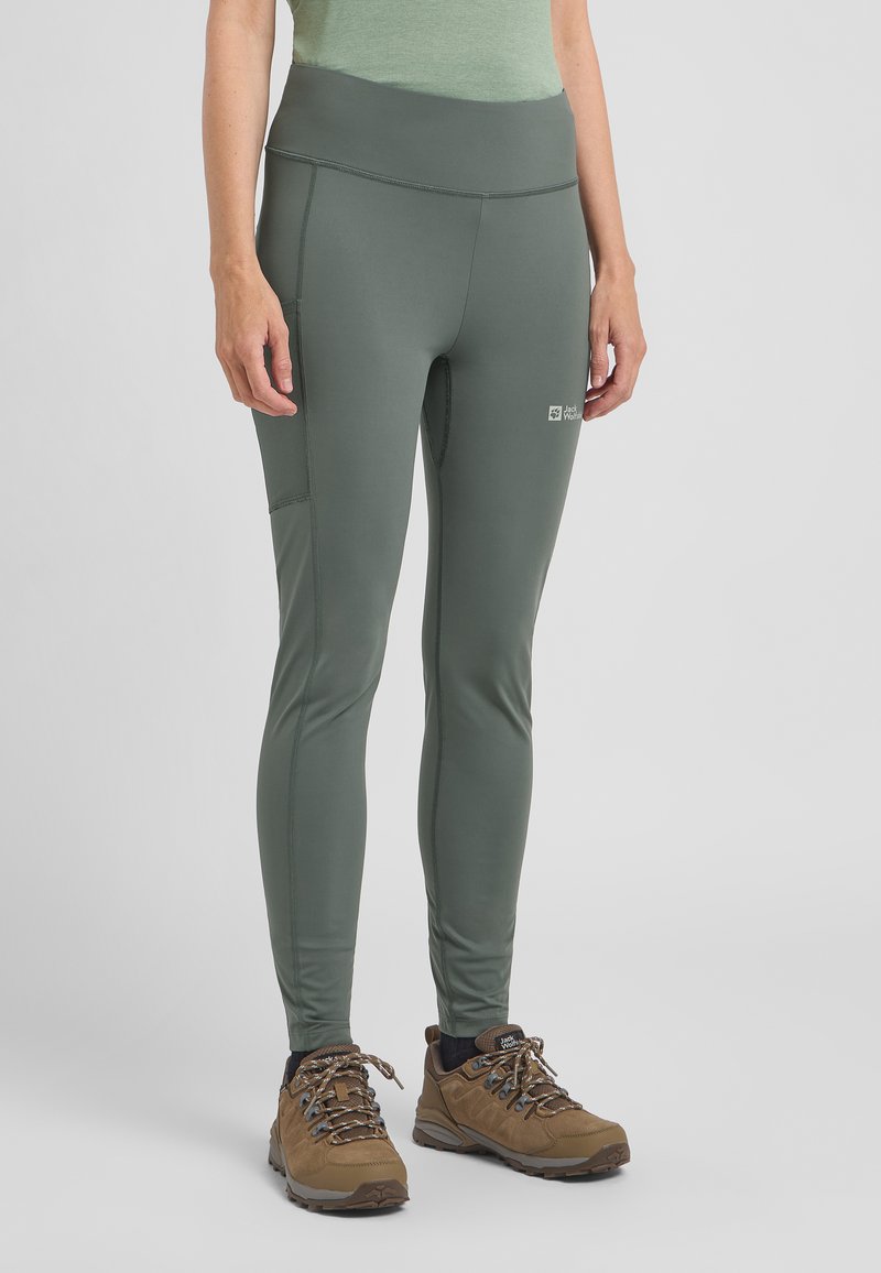 Groene, rekbare legging met een hoge tailleband en zijzak; gecombineerd met bruine wandelschoenen met veters en gestructureerde zolen.