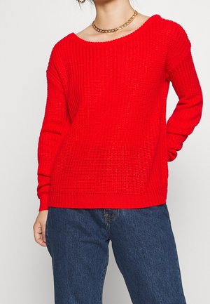 Woman wearing bright red knitted sweater with long sleeves and blue jeans, accessorized with gold chain necklace.