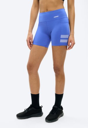 Žena v modrých športových šortkách s vysokým pásom a bočnými vreckami a v čiernych teniskách, stojaca pred jednoduchým svetlým pozadím.