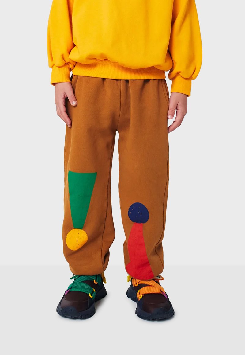 Pantalones de chándal marrones con parches geométricos coloridos en verde, amarillo, azul y rojo, combinados con zapatillas negras con acentos multicolores.