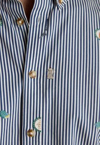 Modrá a biela pruhovaná košeľa s gombíkmi, vyšívanými detailmi vrátane kávovaru a Eiffelovej veže, a rôznymi textúrovanými nášivkami.