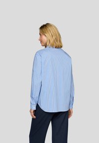 Blue and white striped long-sleeve shirt with a rounded hem, buttoned cuffs, and a relaxed fit, paired with navy textured trousers.