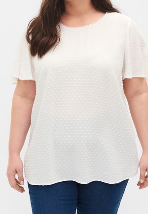 Donna che indossa una camicetta bianca a maniche corte con piccoli puntini in rilievo e jeans blu scuro, vista dalle spalle ai fianchi.