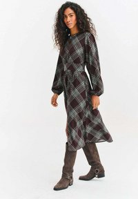 Sukienka w kratę z długim rękawem w ciemnym bordowym i zielonym kolorze, z dopasowaną talią, rozłożystą spódnicą, bocznym rozcięciem, zestawiona z brązowymi, wysokimi kozaczkami do kolan.
